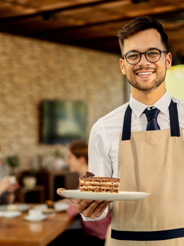 Happy waiter with slice of cake standing in a cafe and looking at camera. There are people in the background.
